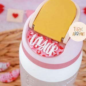 Puede incluir: Un vaso rosa claro y blanco con tapa amarilla, con el nombre "Taylor" en escritura blanca sobre un fondo rosa con estampado de leopardo. El vaso tiene una banda rosa y un logotipo de Three Arrows.