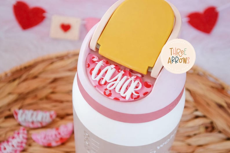 Puede incluir: Un vaso rosa claro y blanco con tapa amarilla, con el nombre "Taylor" en escritura blanca sobre un fondo rosa con estampado de leopardo. El vaso tiene una banda rosa y un logotipo de Three Arrows.