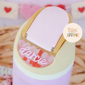 Puede incluir: Un vaso de color p&uacute;rpura claro con una tapa rosa claro y una placa con el nombre decorativa que dice "Darcie". La placa con el nombre tiene un patr&oacute;n a cuadros rojo y blanco. El vaso tiene un asa amarillo claro.