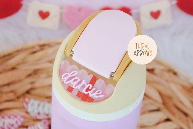 Puede incluir: Un vaso de color p&uacute;rpura claro con una tapa rosa claro y una placa con el nombre decorativa que dice "Darcie". La placa con el nombre tiene un patr&oacute;n a cuadros rojo y blanco. El vaso tiene un asa amarillo claro.