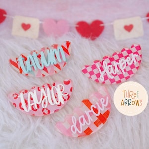 Puede incluir: Cuatro etiquetas de nombre acr&iacute;licas en forma de vieira con varios patrones y nombres. Las etiquetas son rosas, rojas y blancas, con nombres como "Tatum" y "Harper" en cursiva. Una pancarta de corazones y sobres est&aacute; en el fondo.