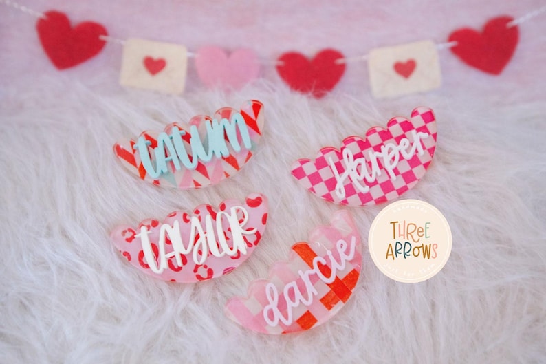 Puede incluir: Cuatro etiquetas de nombre acr&iacute;licas en forma de vieira con varios patrones y nombres. Las etiquetas son rosas, rojas y blancas, con nombres como "Tatum" y "Harper" en cursiva. Una pancarta de corazones y sobres est&aacute; en el fondo.