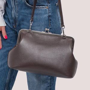 Può includere: Una borsa in pelle marrone con chiusura a scatto e tracolla. La borsa ha una superficie strutturata e una cornice color argento. La borsa è tenuta da una persona che indossa una camicia bianca e jeans blu.