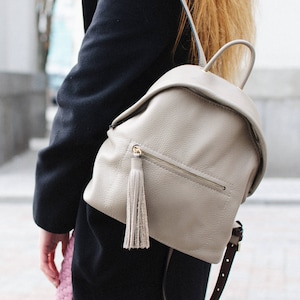 May include: A light beige leather backpack with a tassel zipper pull. The backpack has a flap closure and adjustable straps.