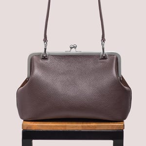 May include: A brown leather handbag with a silver clasp and a single handle. The bag has a textured surface and a rectangular shape. The handle is thin and arched, and the bag is sitting on a wooden surface.