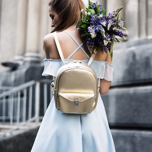 May include: A woman wearing a light blue off-the-shoulder dress with a gold leather backpack. She is holding a bouquet of purple flowers.