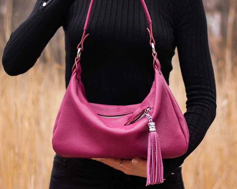 May include: A pink leather handbag with a silver zipper and a tassel. The bag has a shoulder strap and is made of a textured leather.
