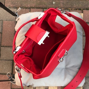 May include: Red leather handbag with a silver clasp and a long adjustable strap. The bag is open and the inside is lined with red fabric.