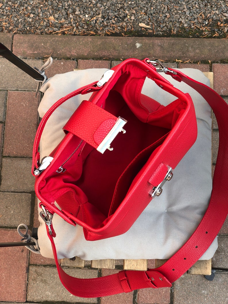 May include: Red leather handbag with a silver clasp and a long adjustable strap. The bag is open and the inside is lined with red fabric.