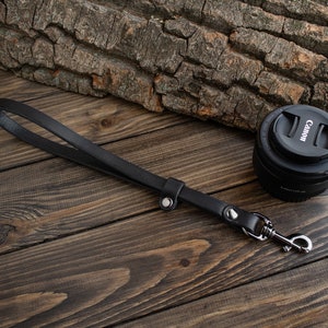 May include: A black leather camera strap with a silver clasp and two silver metal accents. The strap is lying on a wooden surface.