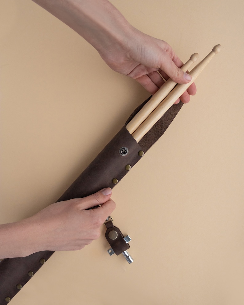 May include: A brown leather drumstick holder with a metal snap closure and a key chain attached. The holder is open and two wooden drumsticks are inside.