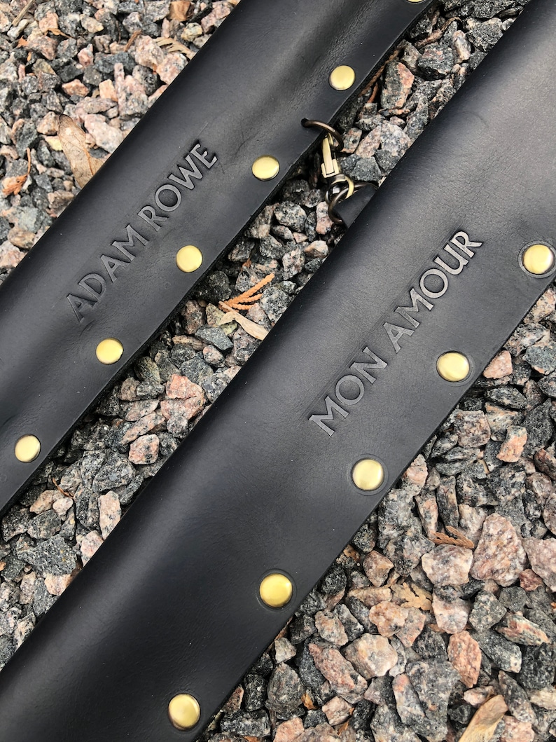 May include: Two black leather straps with gold metal rivets and the text "ADAM ROWE" and "MON AMOUR" embossed on them.