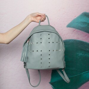 May include: A grey leather backpack with a silver stud design and a tassel. The backpack has a top handle and adjustable straps.
