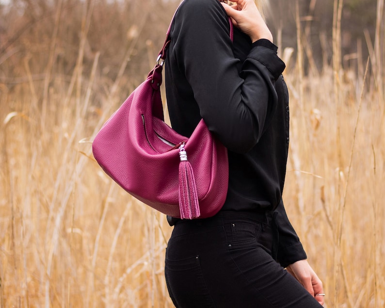 May include: A deep pink leather handbag with a silver tassel and a zipper closure. The bag is worn over the shoulder.