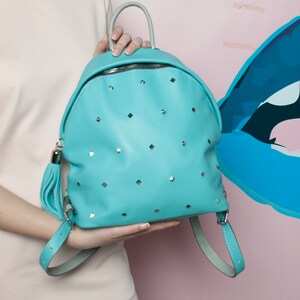 May include: A turquoise blue leather backpack with silver studs and a tassel. The backpack has a rounded shape and adjustable straps.