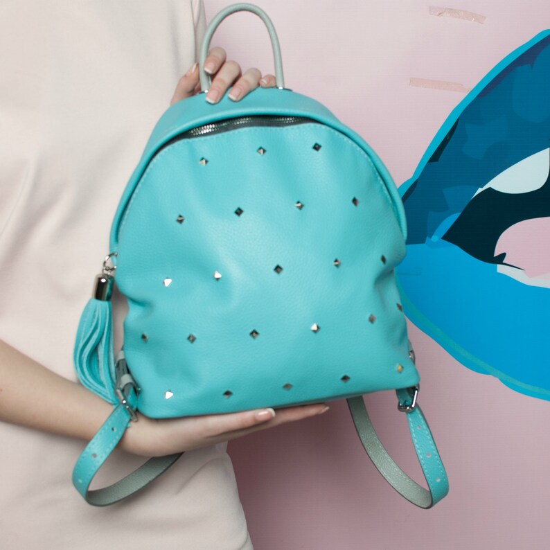May include: A turquoise blue leather backpack with silver studs and a tassel. The backpack has a rounded shape and adjustable straps.