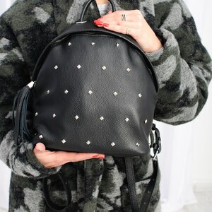 May include: A black leather backpack with silver studs and a tassel. The backpack has a zip closure and adjustable straps.