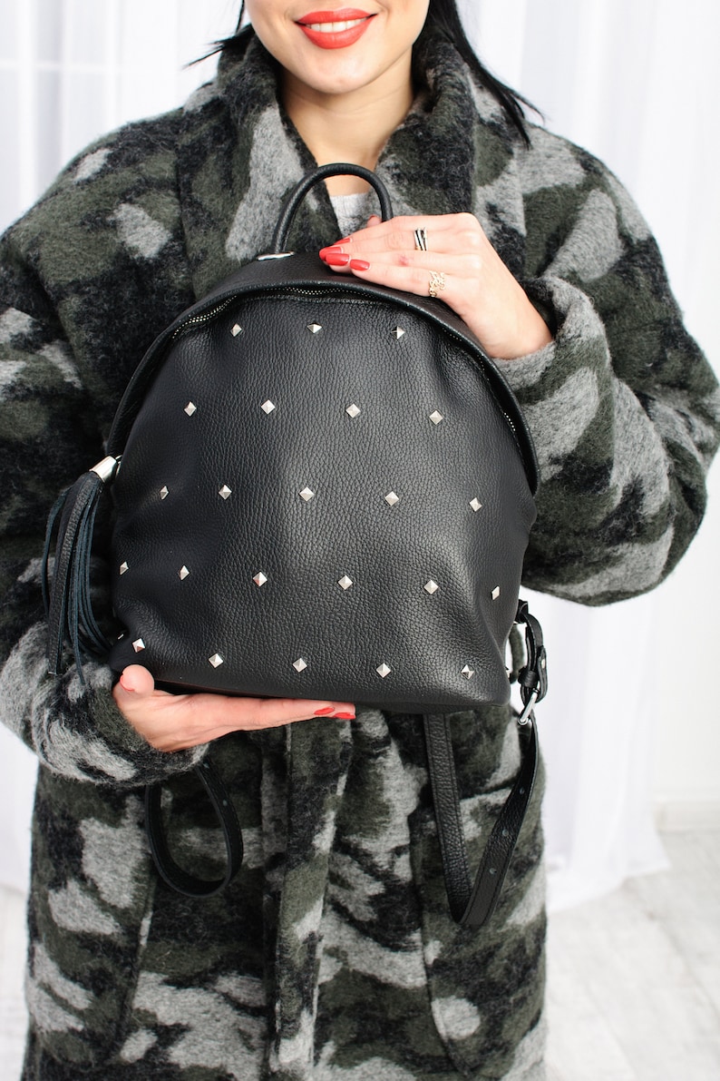 May include: A black leather backpack with silver studs and a tassel. The backpack has a zip closure and adjustable straps.