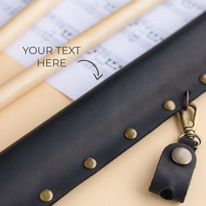 May include: A black leather drumstick case with a metal clasp and a small leather loop with a metal button. The case has brass rivets along the edges.