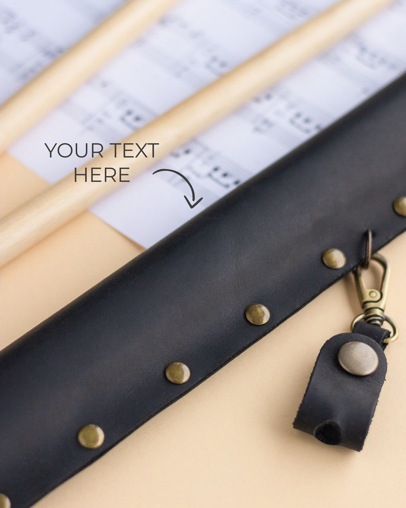 May include: A black leather drumstick case with a metal clasp and a small leather loop with a metal button. The case has brass rivets along the edges.