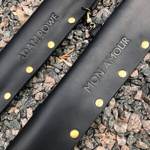 May include: Two black leather straps with gold metal rivets and the text "ADAM ROWE" and "MON AMOUR" embossed on them.