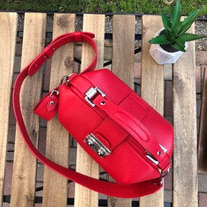 May include: A bright red leather doctor bag with a silver clasp and a long adjustable strap. The bag is sitting on a wooden surface.