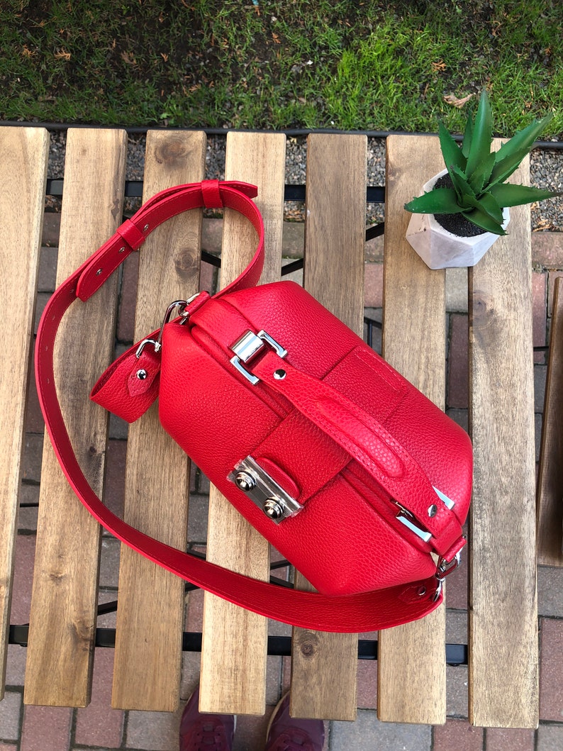 May include: A bright red leather doctor bag with a silver clasp and a long adjustable strap. The bag is sitting on a wooden surface.