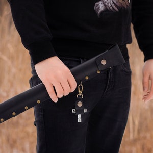 May include: A black leather sheath with a metal clasp and a cross-shaped keychain. The sheath is secured with brass rivets and has a decorative design.