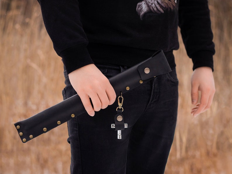 May include: A black leather sheath with a metal clasp and a cross-shaped keychain. The sheath is secured with brass rivets and has a decorative design.