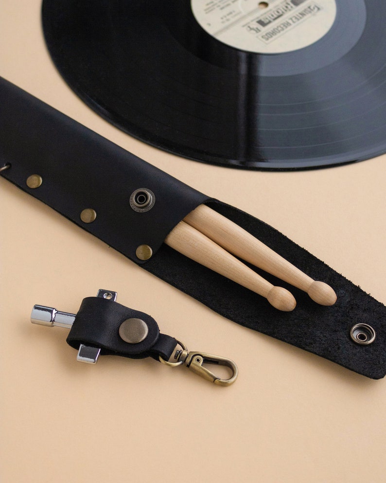 May include: A black leather drumstick holder with two wooden drumsticks inside. The holder has a snap closure and a metal ring for attaching to a belt or bag. A black leather keychain with a metal tool is also pictured.