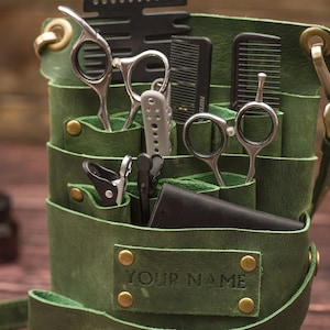 K&ouml;nnte beinhalten: Gr&uuml;ne Ledertasche f&uuml;r Barbierwerkzeuge mit mehreren Taschen und F&auml;chern. Die Tasche ist gef&uuml;llt mit verschiedenen Werkzeugen, darunter Scheren, K&auml;mme und Haarschneider. Die Tasche hat einen Riemen und ein Messing-Namensschild mit der Aufschrift "YOUR NAME".