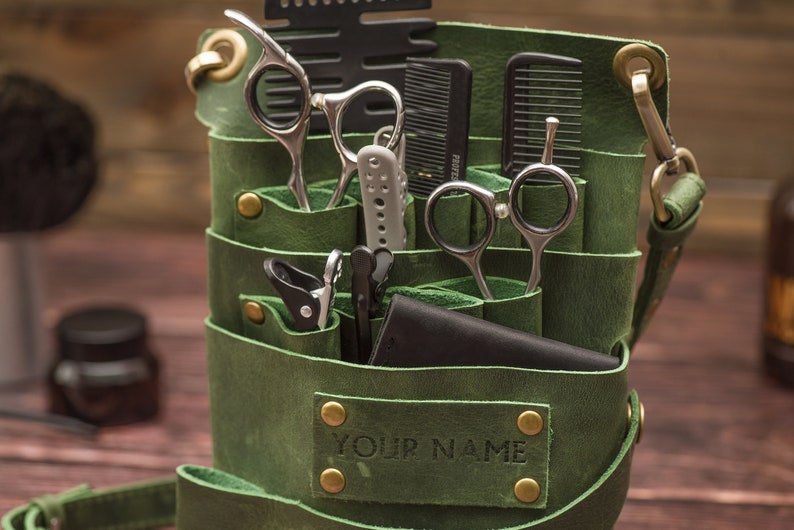K&ouml;nnte beinhalten: Gr&uuml;ne Ledertasche f&uuml;r Barbierwerkzeuge mit mehreren Taschen und F&auml;chern. Die Tasche ist gef&uuml;llt mit verschiedenen Werkzeugen, darunter Scheren, K&auml;mme und Haarschneider. Die Tasche hat einen Riemen und ein Messing-Namensschild mit der Aufschrift "YOUR NAME".