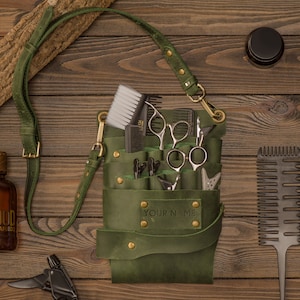 K&ouml;nnte beinhalten: Gr&uuml;ne Ledertasche f&uuml;r Friseurwerkzeuge mit einem Riemen, die Scheren, K&auml;mme und Haarschneider enth&auml;lt. Die Tasche tr&auml;gt den Schriftzug "YOUR NAME" auf der Vorderseite.