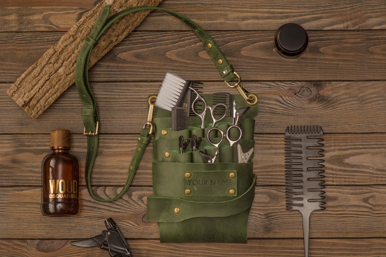 K&ouml;nnte beinhalten: Gr&uuml;ne Ledertasche f&uuml;r Friseurwerkzeuge mit einem Riemen, die Scheren, K&auml;mme und Haarschneider enth&auml;lt. Die Tasche tr&auml;gt den Schriftzug "YOUR NAME" auf der Vorderseite.