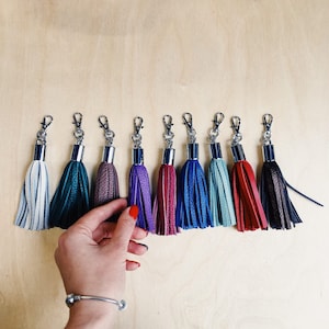 Puede incluir: Diez llaveros de borla de cuero de colores con anillas de plata. Las borlas son en tonos de azul, verde, morado, rojo y marrón.