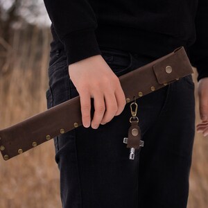 May include: A brown leather sheath with a metal clasp and rivets. The sheath is attached to a key chain with a cross-shaped charm.