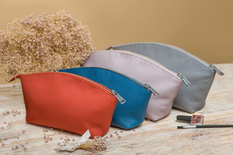 May include: Four zippered pouches in different colors: orange, teal, light gray, and gray. The pouches are made of leather and have a curved shape. They are arranged in a row on a wooden surface.