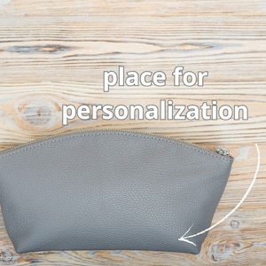 May include: Gray leather cosmetic bag with a zipper closure. The bag has a space for personalization, indicated by an arrow.