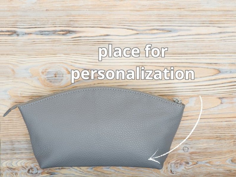 May include: Gray leather cosmetic bag with a zipper closure. The bag has a space for personalization, indicated by an arrow.