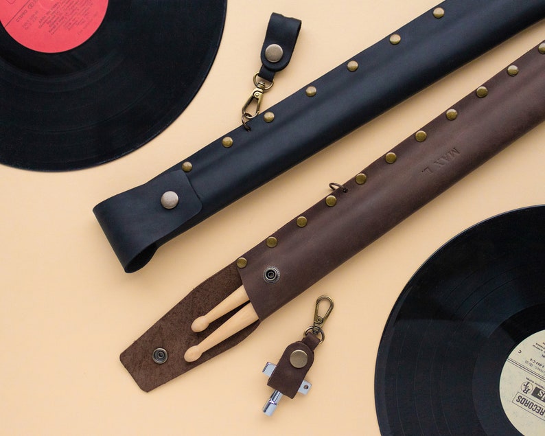 May include: Two leather cases for drumsticks, one black and one brown, with brass rivets. The brown case has the word "MINN" embossed on it. The cases are lying on a beige surface next to two black vinyl records.