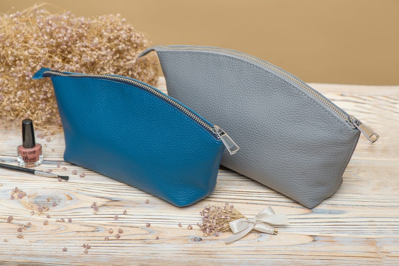 May include: Two leather zippered pouches, one blue and one gray, on a wooden surface.
