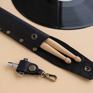 May include: A black leather drumstick holder with two wooden drumsticks inside. The holder has a snap closure and a metal ring for attaching to a belt or bag. A black leather keychain with a metal tool is also pictured.