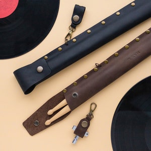 May include: Two leather cases for drumsticks, one black and one brown, with brass rivets. The brown case has the word "MINN" embossed on it. The cases are lying on a beige surface next to two black vinyl records.