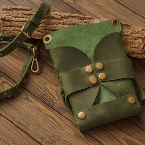 K&ouml;nnte beinhalten: Eine dunkelgr&uuml;ne Ledertasche mit Klappenverschluss und Messing-Akzenten. Die Tasche hat einen langen Riemen und steht auf einer Holzoberfl&auml;che.
