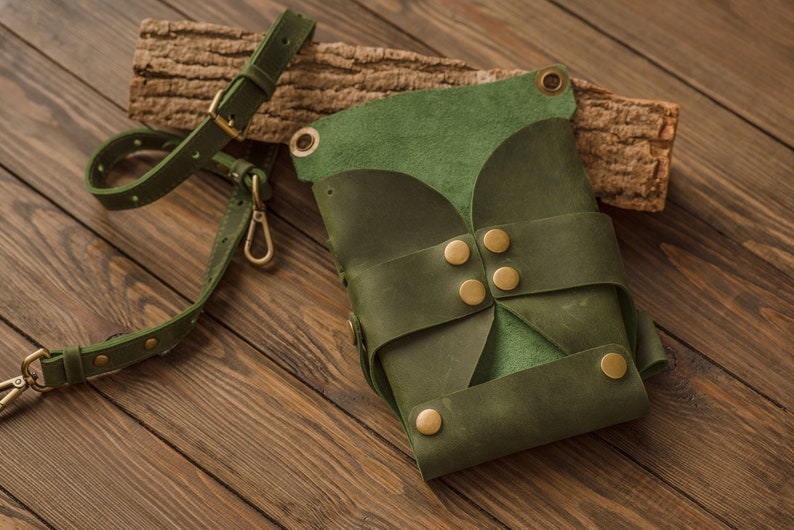 K&ouml;nnte beinhalten: Eine dunkelgr&uuml;ne Ledertasche mit Klappenverschluss und Messing-Akzenten. Die Tasche hat einen langen Riemen und steht auf einer Holzoberfl&auml;che.