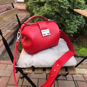 May include: A red leather doctor bag style handbag with a long adjustable strap. The bag has a silver clasp and two silver buttons.