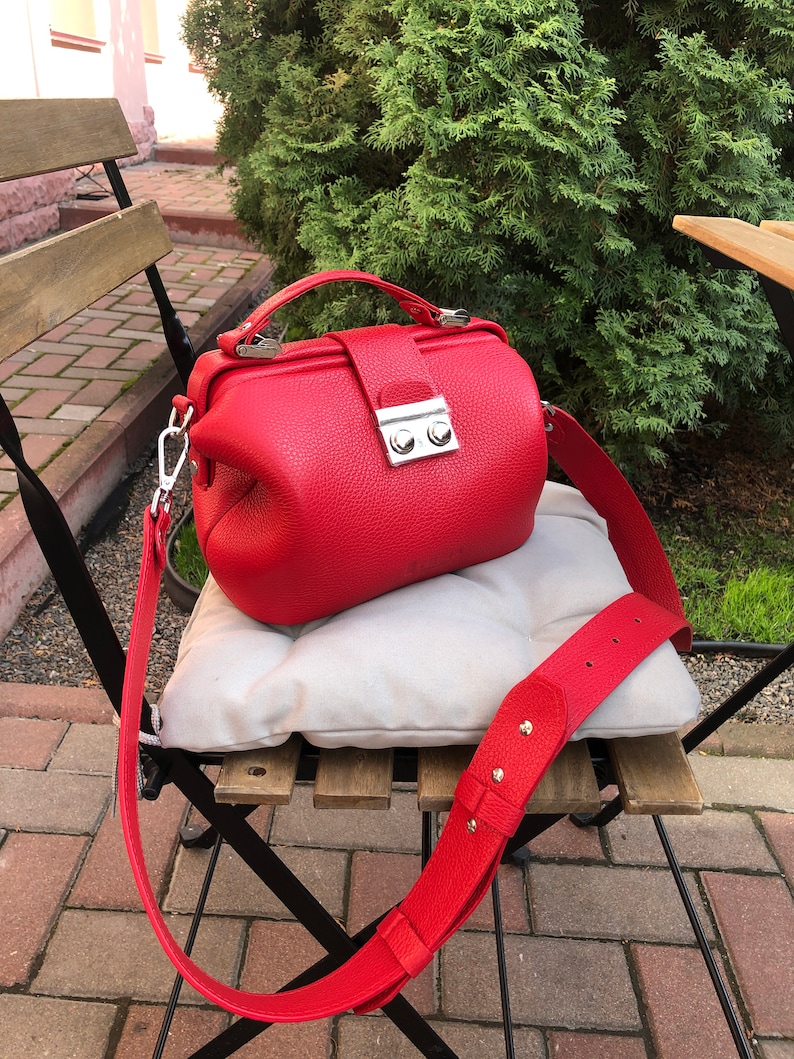 May include: A red leather doctor bag style handbag with a long adjustable strap. The bag has a silver clasp and two silver buttons.