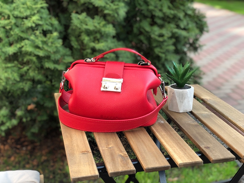 May include: A red leather doctor bag style handbag with a silver clasp and a long strap.