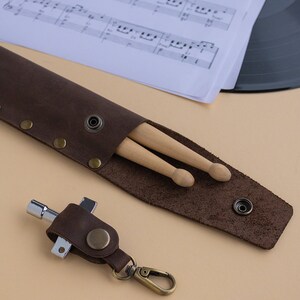 May include: A brown leather drumstick holder with two wooden drumsticks inside. A brown leather keychain with a silver tool is attached to a silver carabiner.