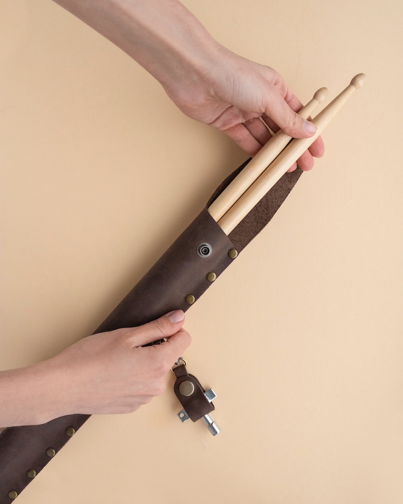 May include: A brown leather drumstick holder with a metal snap closure and a key chain attached. The holder is filled with two wooden drumsticks.